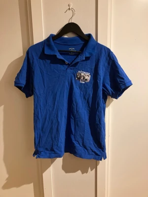 Blå pikétröja Göteborg 1904 - Snygg blå pikétröja med tryck på bröstet där det står 'Göteborg 1904' och en bild på supportrar. Klassisk krage och korta ärmar, perfekt för dig som gillar sportig stil.