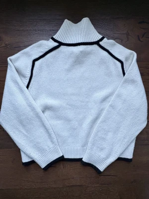 Vit stickad polotröja med svarta detaljer - Snygg vit stickad polotröja med svarta kontrastlinjer längs axlar och hals. Tröjan har ribbstickad hög krage, muddar vid ärmslut och nederkant. 