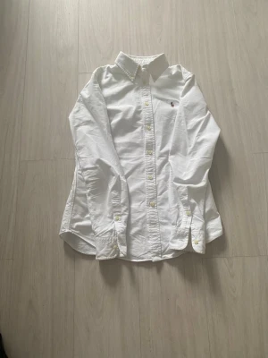 Vit skjorta från Ralph Lauren - Klassisk vit skjorta från Ralph Lauren med broderad logga på bröstet. Skjortan har lång ärm, knappar framtill och krage. Perfekt för en clean och stilren look. Tillverkad i bomull för en skön känsla.