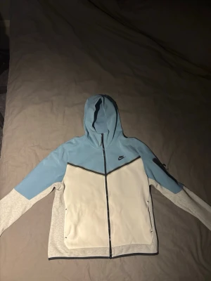 Nike blå och vit hoodie med dragkedja - Nike hoodie i ljusblått och vitt med svarta detaljer. Dragkedja framtill, fickor på sidorna och en liten svart Nike-logga på bröstet. Huva och långa ärmar i kontrasterande färger. Tillverkad i mjukt bomullsmaterial för en skön känsla.