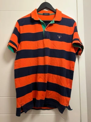 Randig pikétröja från Gant - Snygg pikétröja från Gant med breda orange och marinblå ränder. Klassisk krage med grön insida och korta ärmar. Gant-logga broderad på bröstet. Tillverkad i mjuk bomull, perfekt för en avslappnad stil.