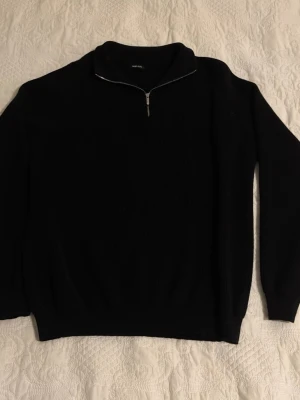 Svart stickad halvzip från tour&tate - Svart stickad tröja från tour&tate med halvzip och hög krage. Tröjan har ribbad struktur, långa ärmar och en stilren metallblixtlås framtill. Perfekt för lager på lager och enkel att matcha med olika outfits.