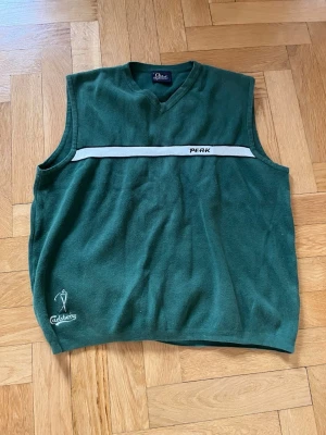 Grön stickad väst från Peak - Grön stickad golf-väst från Peak performance med vit och svart rand över bröstet och broderad logga. V-ringad modell med Carlsberg-brodyr nertill. Står storlek Large men passar nog de flesta storlekar. Bra skick o inga fläckar eller skavanker.                                                                Längd: 62 cmBredd: 41 cm