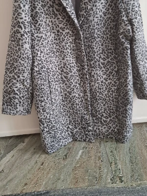 Leopardmönstrad kappa från Sora - Stilren leopardmönstrad kappa från Sora by jbc i storlek 42. Kappan har två knappar framtill, sidofickor och är fodrad. Färgen är grå med svarta fläckar och materialet känns mjukt, perfekt för att sticka ut i mängden.