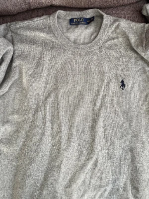 Grå stickad tröja från Polo Ralph Lauren - Snygg grå stickad tröja från Polo Ralph Lauren med klassisk rund hals och diskret broderad logga på bröstet. Tröjan har långa ärmar och ribbade muddar vid ärmslut och nederkant. Perfekt för dig som gillar stilren och enkel design.
