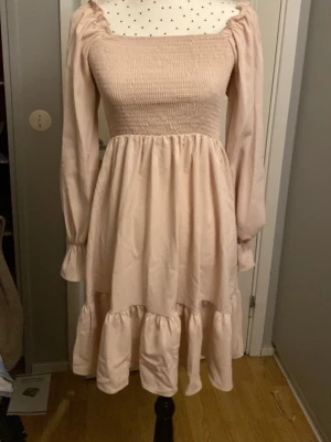 Ljusrosa volangklänning med långa ärmar - Supersöt ljusrosa klänning med smockad överdel, volangkant nertill och långa ärmar med resår vid handlederna. Klänningen kan bäras både på axlarna och offshoulder. 