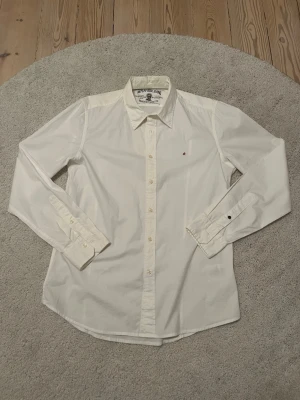 Replay skjorta - Klassisk vit skjorta från Replay Blue Jeans med diskret röd broderad 'R' på bröstet. Skjortan har lång ärm, knappar framtill och på manschetterna samt en snygg krage. Perfekt till en clean och stilren look.