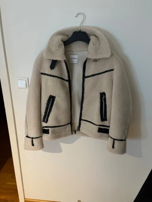 Beige teddyjacka från Bershka - Snygg beige teddyjacka från Bershka med svarta detaljer och dragkedjor. Jackan har hög krage med spänne, två framfickor med dragkedja och markerade svarta sömmar. Perfekt för att hålla värmen och se trendig ut under kyliga dagar.