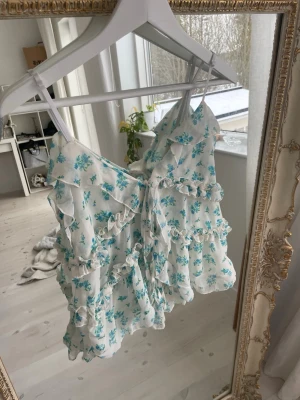 Blommig volangtopp med knyt - Supersöt vit topp med blått blommönster och volanger. Toppen har smala axelband, v-ringning och flera knyt framtill. Luftig och flowy modell som ger en romantisk vibe, perfekt för varma dagar. Original pris är 378 kr.