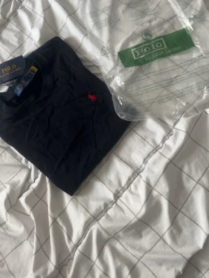 Svart t-shirt från Polo Ralph Lauren - Klassisk svart t-shirt från Polo Ralph Lauren med den ikoniska röda logon broderad på bröstet. T-shirten har rund hals, korta ärmar och är gjord i mjuk bomull för en skön känsla. Perfekt basplagg med snygg passform.