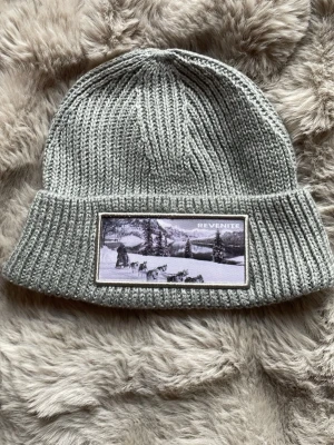 Grå stickad mössa från Revenite - Snygg grå ribbstickad mössa från Revenite med uppvikt kant. Framsidan har en patch med svartvit vinterbild och Revenite-logga. Perfekt accessoar för kalla dagar och enkel att matcha med olika outfits.