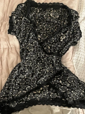 Svart spetsmönstrad topp från Promod - Snygg svart topp från Promod med vit blommig spets och romantiskt rosmönster. Toppen har korta ärmar, omlott-skärning och dekorativ spetskant vid hals och nederkant. Perfekt för dig som gillar en feminin och trendig stil. Passar mig Som är xs