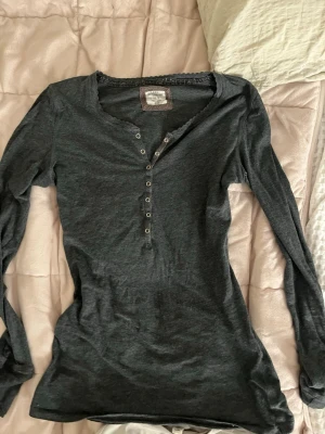 Mörkgrå långärmad topp med knappar - Mörkgrå långärmad topp med knappdetaljer framtill och rundad halsringning. Tillverkad i mjukt bomullsmaterial som känns skönt mot huden. Enkel och stilren design som passar till det mesta. Litet slitmärke på ena armbågen och liten fläck mitt på magen.#y2k #elanagillbert.