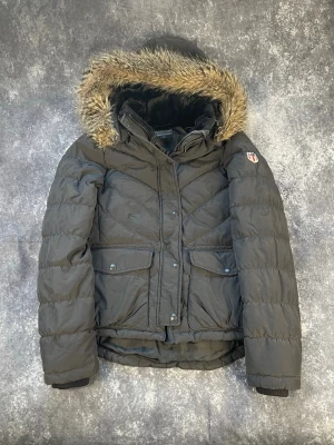 Svart dunjacka med fuskpäls från Ralph Lauren - Snygg svart dunjacka från RL med avtagbar huva och fuskpäls. Jackan har två stora fickor framtill, ribbade muddar och ett broderat RL-märke på ärmen. Perfekt för kalla vinterdagar och riktigt varm tack vare dunfyllningen.