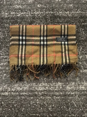 Burberrys rutig halsduk i lammull - Klassisk rutig halsduk från Burberrys i beige, svart, vitt och rött. Tillverkad i 100% lammull och har fransar i ändarna. Den ikoniska rutiga designen gör den superenkel att matcha med olika outfits.