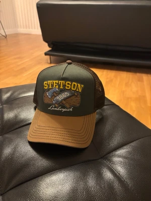 Stetson Lumberjack truckerkeps - Snygg truckerkeps från Stetson med grönt tyg framtill, brunt skärm och mesh i mörkbrunt bak. Broderad logga och motiv med texten 'Lumberjack 1865' i gult och vitt. Perfekt för dig som gillar retrostil och streetwear. Samt helt ny aldrig använd. Nypris 499 mitt pris 260