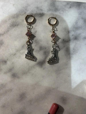 Guldörhängen med skridskohänge - Unika örhängen med guldfärgade ringar, glittrande pärldetaljer i rosa och små skridskoformade hängen i silverfärg. Perfekta för dig som vill sticka ut med en sportig och lekfull stil. Materialet är metall och plast.