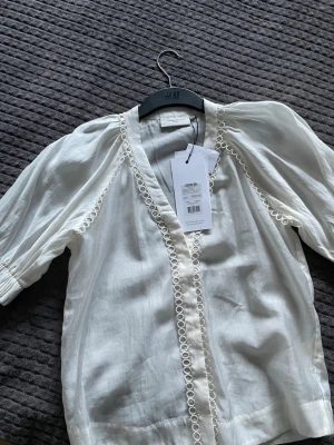 Vit blus från Copenhagen Muse - Stilren vit blus från Copenhagen Muse i luftig bomull med dekorativa broderade detaljer längs kanterna. Blusen har v-ringning och trekvartsärm med lätt puffad axel för en trendig look. Perfekt att matcha med jeans eller kjol.