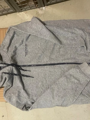 Grå hoodie från Race Marine - Snygg grå hoodie från Race Marine med dragkedja framtill och justerbar huva med svarta snören. Tillverkad i mjukt sweatshirtmaterial och har fickor på sidorna. Diskret logga på bröstet och en liten detalj på ärmen. Perfekt för en avslappnad stil.