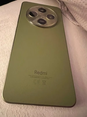 Grön Redmi Android-telefon - Snygg grön Redmi Android-telefon med stor rund kameramodul på baksidan och flera linser. Baksidan har en matt finish och telefonen har en modern, rektangulär form med rundade hörn. Perfekt för dig som gillar stilren design och vill ha en kraftfull kamera.