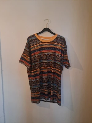 Mönstrad t-shirt från Hoodie Lab - Säljer en färgglad t-shirt från Hoodie Lab med tribal-inspirerat mönster i orange, blått, svart och gult. T-shirten har rund halsringning med orange kant och korta ärmar. Perfekt för dig som gillar unika prints och streetwear vibes. Liten i storlek som en xl