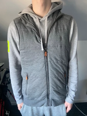 Grå Ralph Lauren väst - Snygg grå dunväst från Polo Ralph Lauren med dragkedja framtill och två fickor med bruna detaljer. Västen har en diskret logga på bröstet och ribbade muddar nedtill. Perfekt att slänga över en hoodie för en chill look. Fint skick, passar dig mellan 180-185.