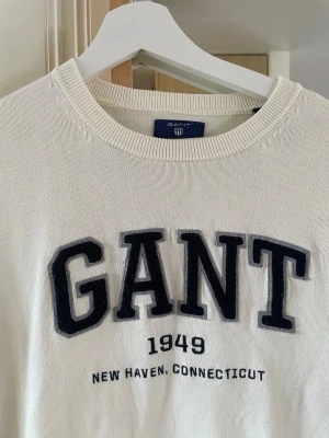 Vit college-tröja från GANT - Vit långärmad college-tröja från GANT med stor svart broderad logga och texten '1949 New Haven, Connecticut' på bröstet. Tröjan har rund halsringning och är gjord i mjukt material som känns skönt mot huden. Perfekt för en avslappnad och stilren look. Herr!  Stl M 