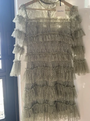 Grön spetsklänning från Malina - Superfin grön kort klänning från Malina i spets med volangdetaljer över hela klänningen. Aldrig använd och har lappen kvar! Perfekt till lite festliga tillfällen.
