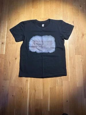 Svart Maison Margiela t-shirt - Svart t-shirt från Maison Margiela med siffror-tryck på bröstet i en vit, tvättad effekt. T-shirten har klassisk passform och korta ärmar. Perfekt för dig som gillar streetwear och vill sticka ut med en unik design. Storlek S men passar in mer på XS.