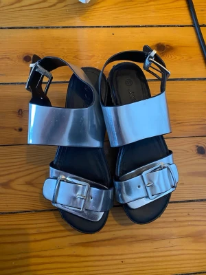 Chunky silvriga sandaler med spänne - Chunky sandaler i metallic silver med breda remmar och stora spännen. Sandalerna har en rejäl plattformsula och klack, samt öppen tå. Materialet är syntet och sulan är svart. Märke Monki.