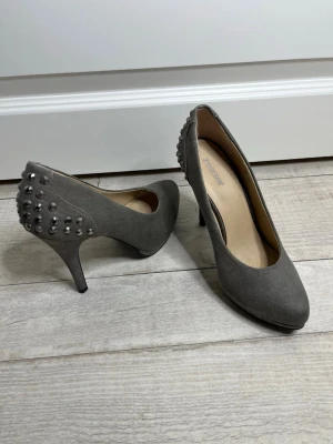 Grå pumps med nitar från Graceland - Snygga grå pumps från Graceland i storlek 36 men passar också 37, skorna är med rundad tå och klädd klack. Baksidan är dekorerad med silvriga nitar som ger en cool touch. Ovandelen är i mockaimitation och innersulan är beige. Perfekta för dig som vill sticka ut lite extra.