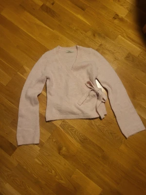 Ljusrosa omlottkofta från Gina Tricot - Supersöt ljusrosa stickad omlottkofta från Gina Tricot med knytband i midjan. Koftan har lång ärm och mjuk, fluffig känsla. Perfekt att slänga över en topp för en chill och trendig look.