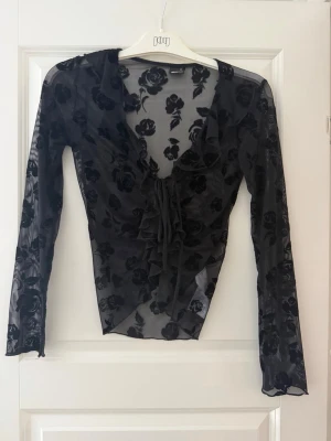 Svart meshblus med blommönster - Säljer en transparent svart meshblus från Gina med blommigt mönster och långa ärmar. Blusen har v-ringning, volangdetaljer framtill och knyt i midjan. Endast använd 1 gång