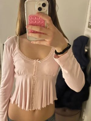 Ljusrosa långärmad croppad topp - Supersöt ljusrosa långärmad topp med volangkant och liten rosett framtill. Toppen är croppad och har spetsdetaljer längs urringningen och framtill. 