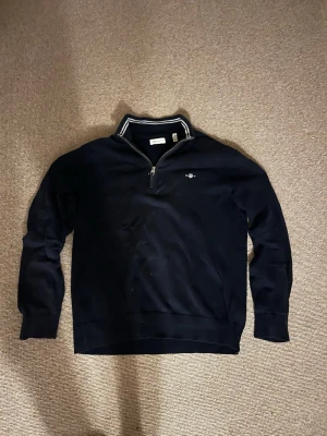 Marinblå halvzip från Gant - Snygg marinblå tröja från Gant med halvzip och ståkrage. Tillverkad i mjukt material och har ribbade muddar vid ärmslut och nederkant. Hör av dig vid minsta fundering!😊