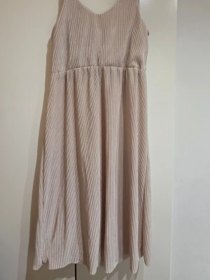 Beige klänning med smala ränder - Säljer en beige långklänning  med smala vertikala ränder. Klänningen har ärmlös design och v-ringad hals. Perfekt för dig som gillar en avslappnad och stilren look.