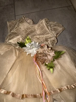 Champagnefärgad klänning med blommor - Söt champagnefärgad klänning med korta ärmar och volangkjol i tyll. Livet har ett diskret mönster och midjan pryds av dekorativa blommor och band i pastellfärger. Perfekt för speciella tillfällen när du vill känna dig extra fin.