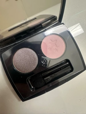 Lancôme duo ögonskugga rosa & lila/grå - Snygg duo ögonskugga från Lancôme i en svart kompakt dosa med spegel. Ena skuggan är skimrande lila/grå och den andra är matt ljusrosa med stjärnmönster. Medföljer en liten applikator. Perfekt för att skapa olika ögonlooks