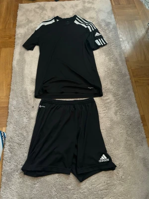 Svart Adidas träningsset herr - Säljer ett svart träningsset från Adidas med t-shirt och shorts. T-shirten har ett litet slitage märke där bak som man ser på en av bilderna men absolut inget stort. Annars är kitet i relativt fint skick. 
