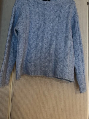 Ljusblå kabelstickad tröja - Mysig ljusblå stickad tröja med klassiskt kabelstickat mönster. Tröjan har rund halsringning, långa ärmar och ribbade muddar vid ärmslut och nederkant. Perfekt för kyliga dagar och enkel att matcha med jeans eller kjol ifrån lager 157
