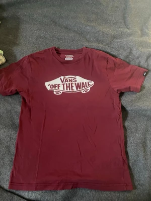 Vinröd Vans t-shirt med tryck - Säljer en vinröd t-shirt från Vans med det klassiska 'Off The Wall'-trycket i vitt på bröstet. T-shirten har rund hals och korta ärmar, perfekt för en avslappnad streetwear-look.