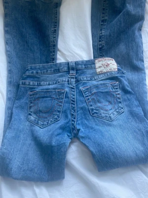 Blå bootcut jeans från True Religion - Säljer ett par klassiska blå bootcut jeans från True Religion med ikoniska bakfickor och kontrastsömmar. Jeansen har låg midja. Perfekta för dig som gillar en avslappnad men snygg stil. Men det är slitets längst ner på jeansen!!