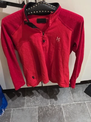 Röd fleece från Haglöfs med halv dragkedja - Röd fleece från Haglöfs med halv dragkedja och hög krage. Tröjan har lång ärm och är tillverkad i mjukt fleecematerial. Perfekt för lager-på-lager och har en liten Haglöfs-logga broderad på bröstet.