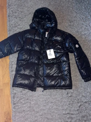 Svart glansig dunjacka från Moncler - Snygg svart dunjacka från Moncler med glansigt yttertyg och klassisk puffer-design. Jackan har huva, dragkedja framtill och Moncler-logga på ärmen. Perfekt för kalla vinterdagar och riktigt trendig look.
