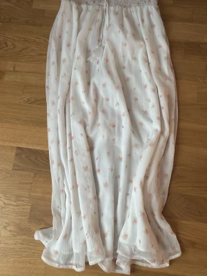 Vit långkjol med små blommor - Luftig vit långkjol med små rosa blommor och smockad midja med snörning och tofsar. Kjolen har ett romantiskt och somrigt uttryck, perfekt för varma dagar. Tunt, lätt tyg som ger ett flowy fall. Storlek 158/164!!!