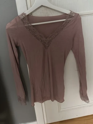 Ljuslila långärmad topp med spets - Söt ljuslila långärmad topp med v-ringning och fina spetsdetaljer vid halsringning och ärmslut. Perfekt att styla med jeans eller kjol för en trendig look.