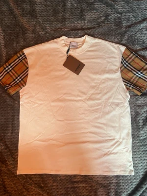 Vit t-shirt med rutiga ärmar Burberry - Snygg vit t-shirt från Burberry med klassiska rutiga ärmar i beige, svart, rött och vitt. T-shirten har en rak passform och är tillverkad i mjuk bomull. Perfekt för dig som vill sticka ut med en ikonisk detalj.