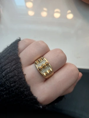 Bred guldfärgad ring med stenar - Cool bred ring i guldfärg med glittrande stenar och dekorativa nitar. Ringen har en chunky design och detaljer i både guld och silverton. Perfekt för dig som gillar statement-smycken och vill sticka ut.