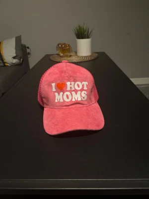 Rosa keps med text I ❤️ HOT MOMS - Cool rosa keps med vit broderad texten 'I ❤️ HOT MOMS' framtill. Kepsen har böjd skärm och mesh-tyg baktill för extra ventilation. Perfekt accessoar för att sticka ut och visa attityd.
