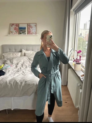 Ljusgrön trenchcoat med skärp - Snygg ljusgrön trenchcoat med bred krage och knytskärp i midjan. Jackan har långa ärmar och en klassisk, rak passform. Perfekt för dig som vill ha en stilren och trendig look. Materialet ser ut att vara mjukt tyg, vilket ger en avslappnad känsla.